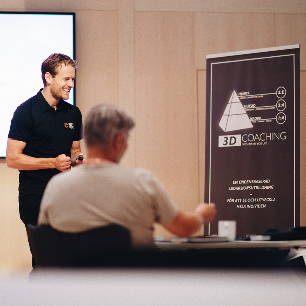 Leende person står framför en annan som sitter vid ett bord och antecknar 